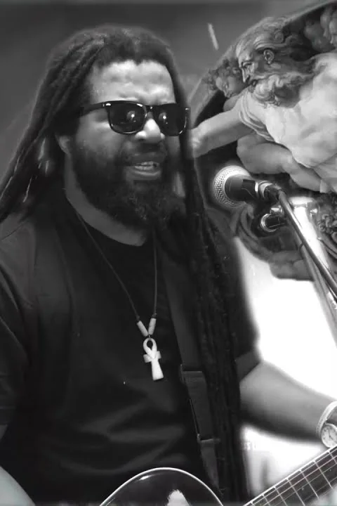 Mutti Lewis in black and white, singing into a microphone with an acoustic guitar, wearing sunglasses and an ankh on a cord